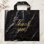 A black plastic 'Thank You' bag with gold script lies flat on a white marble surface with eucalyptus leaves.