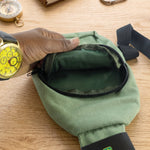A hand opens the main zipper compartment of the olive green canvas sling bag, revealing a dark green interior.