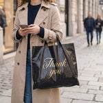 A person in a trench coat holds a black plastic 'Thank You' bag while looking at their phone on a city street.