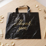 A black plastic 'Thank You' bag with gold script is laid flat on a wooden surface with scattered rose petals.