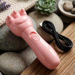 A pink bear paw massager and its black USB cable rest on a wooden surface with decorative stones and plants.