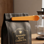 A black coffee bag is sealed with an orange plastic bag clip on a wooden table.