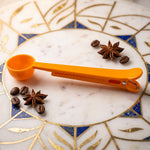 An orange plastic scoop and clip rests on a decorative white and blue marble surface, surrounded by coffee beans and star