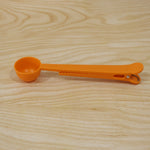 A simple orange plastic measuring spoon with a clip is positioned on a light brown wooden surface.