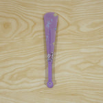 A closed purple folding hand fan with a subtle cat and bow pattern lies vertically on a light wooden surface.