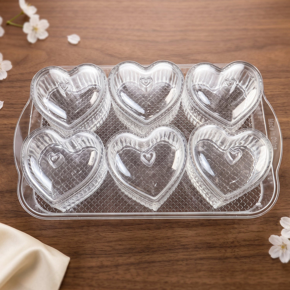 Six clear heart-shaped containers with ribbed sides and matching lids are arranged on a transparent rectangular tray,