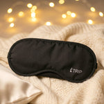 A black sleeping eye mask with the "iTRIP" logo lies on a cream cable-knit blanket, with warm fairy lights in the background.