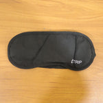 A black sleeping eye mask with the "iTRIP" logo is laid flat on a light brown wooden surface.