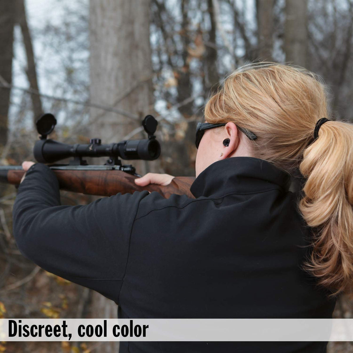 A woman with blonde hair in a ponytail aims a rifle, wearing a black earplug in her right ear.