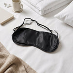 A black silk satin sleep mask lying on a white bed with a book, mug, and white bedding.
