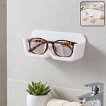 White plastic wall-mounted eyewear holder with tortoise-shell glasses, above a sink with a small green plant