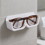 White plastic wall-mounted eyewear holder with tortoise-shell glasses, positioned on a tiled wall next to a mirror.