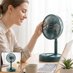 A woman adjusts a dark green portable fan on a wooden desk with a laptop and coffee mug.