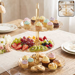 A three-tier amber acrylic serving stand holds colorful cupcakes, fresh fruit, and cookies on a wooden table.