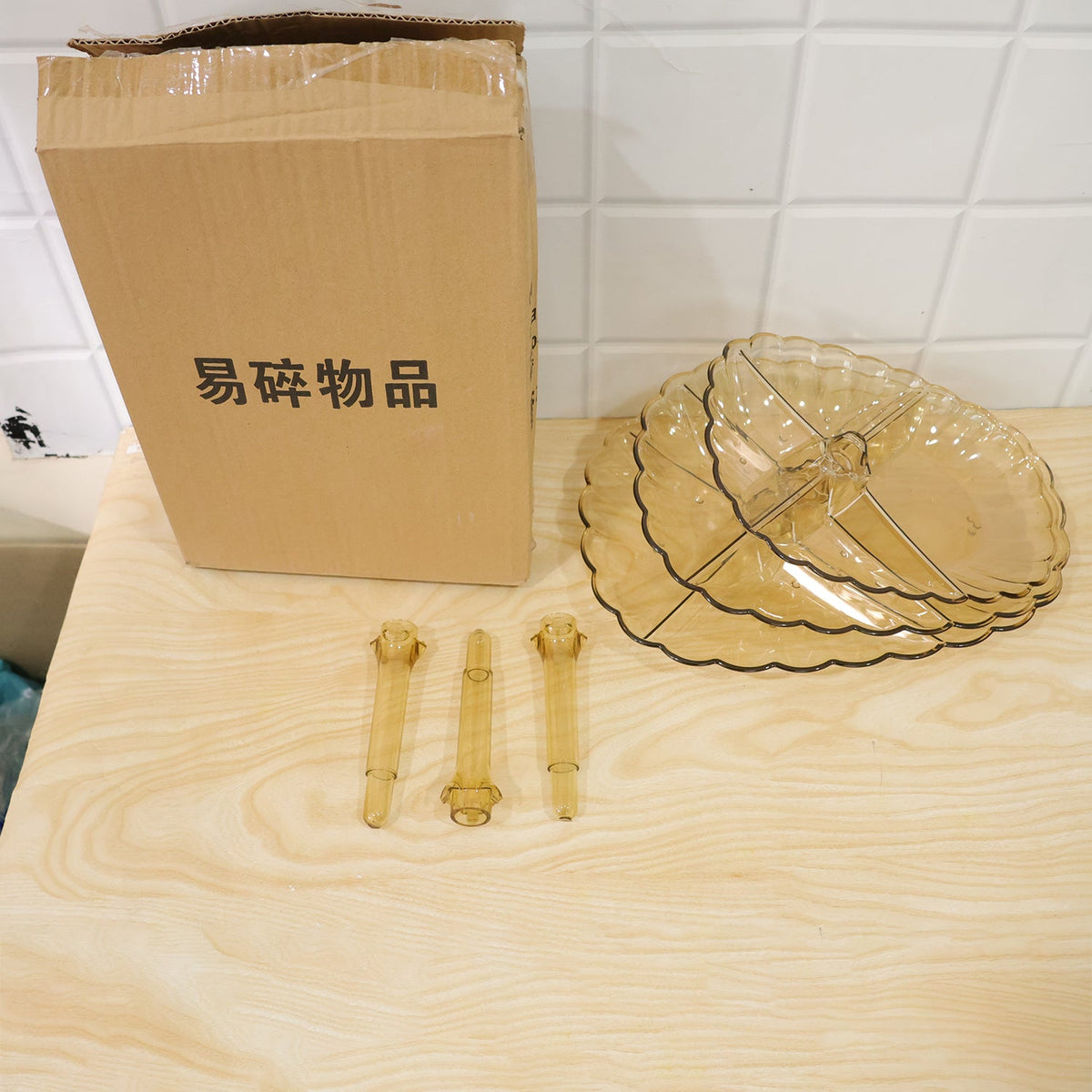 Disassembled amber acrylic serving stand components, including plates and rods, are next to a cardboard box.
