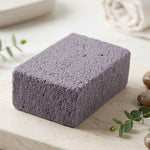 A single gray cleaning block with a porous texture resting on a light marble surface with green leaves and pebbles.