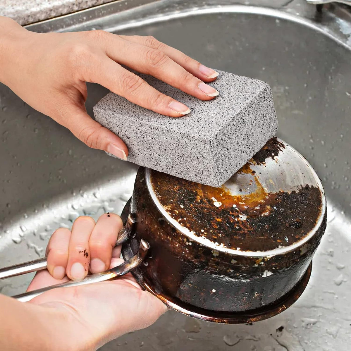 A hand holding a gray cleaning block, scrubbing the burnt exterior of a metal pot in a stainless steel sink.