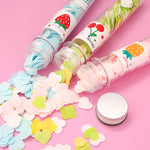Three clear tubes of colorful flower-shaped paper soap strips are spilling out onto a pink surface, with a silver cap nearby.