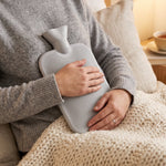 A person in a grey sweater holds a grey ribbed hot water bottle to their chest, with a cream knitted blanket on their lap.