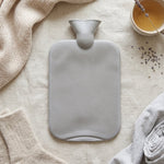 A grey ribbed hot water bottle is centered on a light fabric, surrounded by a knitted scarf, socks, folded fabric, and a cup
