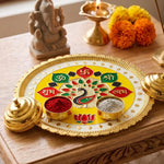 A yellow and gold puja thali with peacock design, small bowls of red and grey powder, and a Ganesha statue in the background.
