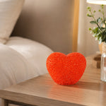 A red heart-shaped LED night lamp rests on a wooden nightstand beside a small plant in a clear vase.
