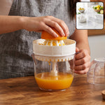 A person's hands squeezing an orange half onto a white plastic reamer, collecting juice in a clear cup.