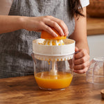 A person's hands squeezing an orange half onto a white plastic reamer, collecting juice in a clear cup, on a wooden surface.
