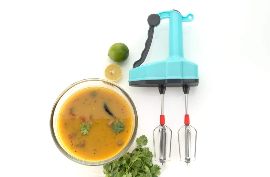 A turquoise manual hand blender is displayed next to a bowl of yellow soup, fresh cilantro, and lime wedges on a white