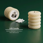 Two cream ribbed LED candles, one showing the battery compartment, on a dark green surface.