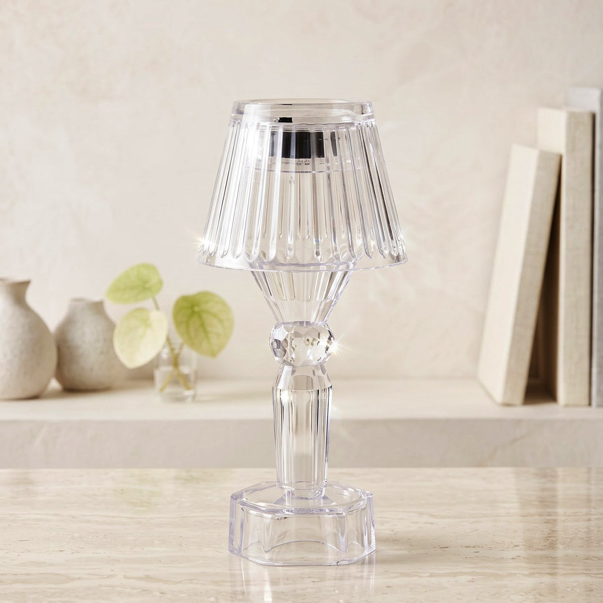A single clear acrylic miniature lamp with a ribbed shade sits on a marble surface, with blurred books and vases in the