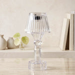A single clear acrylic miniature lamp with a ribbed shade sits on a marble surface, with blurred books and vases in the