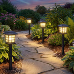 Four black solar lanterns cast a warm glow along a stone path in a vibrant, green garden at twilight.
