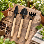 Three miniature gardening tools with wooden handles rest on a rustic wooden table surrounded by potted plants.
