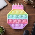 A closed pastel hexagon pop it fidget notebook on a wooden desk with a laptop, coffee cup, and plant.