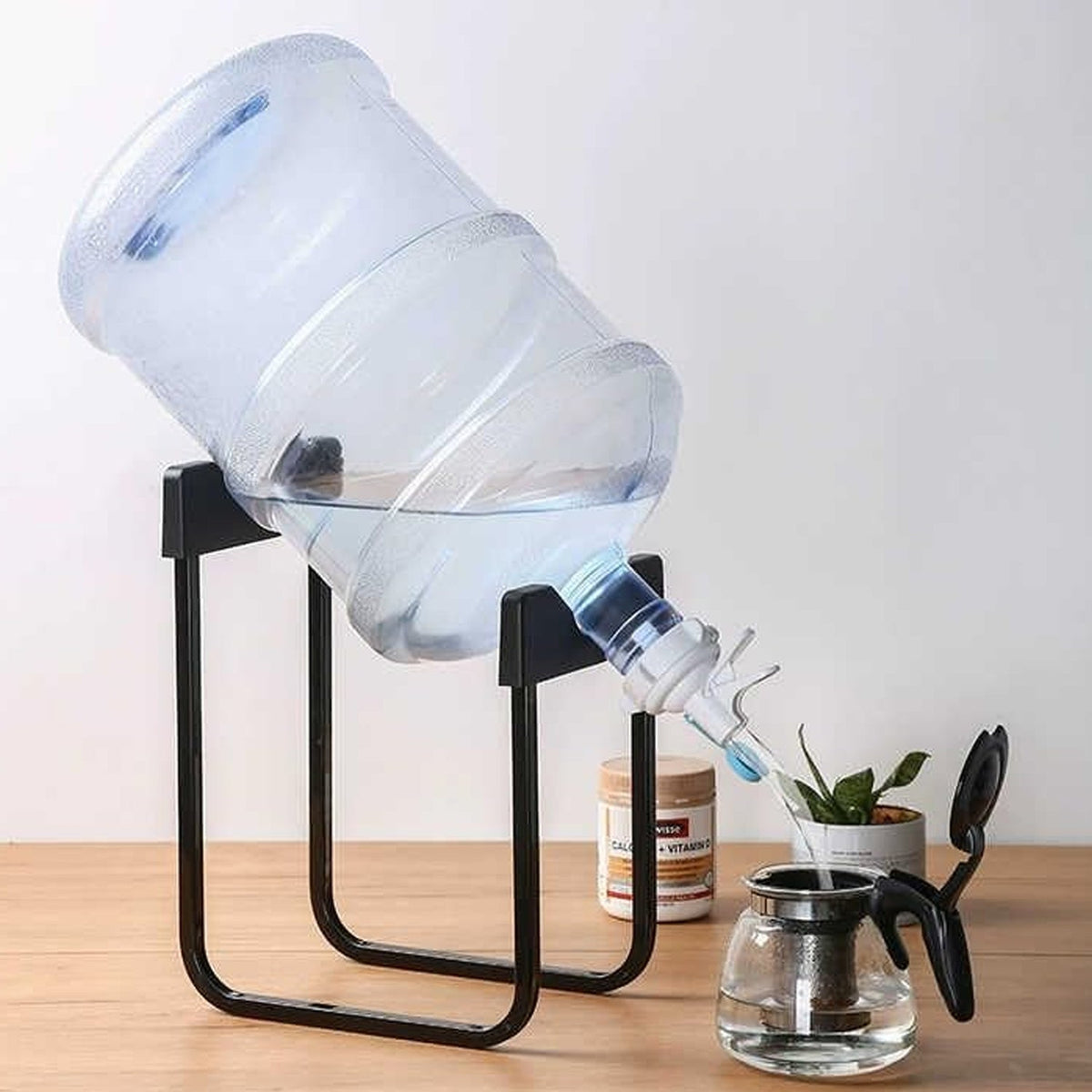 A large clear water bottle sits tilted on a black metal stand, dispensing water into a glass teapot on a wooden surface.