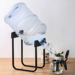 A large clear water bottle sits tilted on a black metal stand, dispensing water into a glass teapot on a wooden surface.