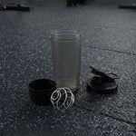 A disassembled grey and black shaker bottle with a metal whisk is on a dark gym floor.