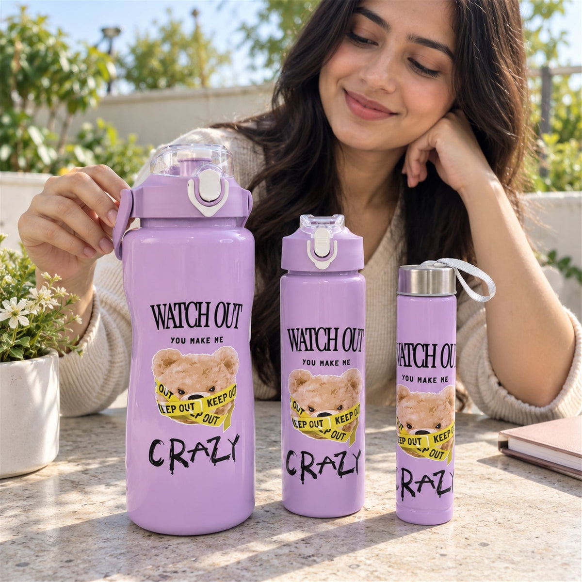 A person gently touching the largest purple water bottle, part of a set of three with a teddy bear design, outdoors.