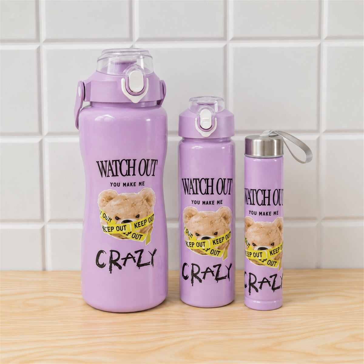 Three purple water bottles with a teddy bear design, in varying sizes, on a wooden surface against a white tiled wall.