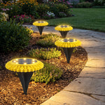 Four yellow and black solar ground lights illuminate a garden path at dusk, with green foliage and a stone walkway.