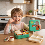 A child with light brown hair smiles while eating from a green dinosaur lunchbox filled with a sandwich, fruit, and