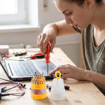A person uses a red and black screwdriver to work on a laptop, with the yellow and clear screwdriver set on a wooden table.