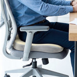 A person sitting on a grey office chair with a beige and grey ergonomic seat cushion.