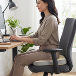 A woman sitting at a desk on a grey office chair with a beige and grey ergonomic seat cushion, typing on a laptop.