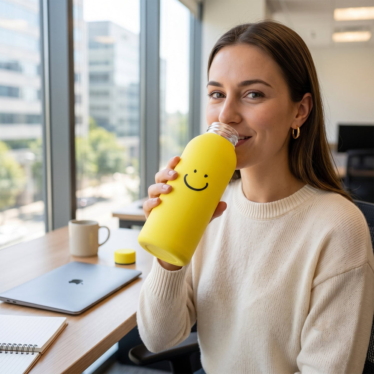 A woman smiling and drinking from a yellow glass water bottle with a smiley face at an office desk.