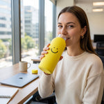 A woman smiling and drinking from a yellow glass water bottle with a smiley face at an office desk.