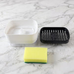 The white soap dispenser base, black perforated top, and yellow sponge are separated on a white marble surface with water