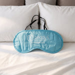 A light blue sleep mask with a white logo and black elastic strap rests on a neatly made white bed with pillows.