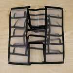 A white mesh and black fabric 6-section drawer organizer is shown folded flat on a light wooden surface.